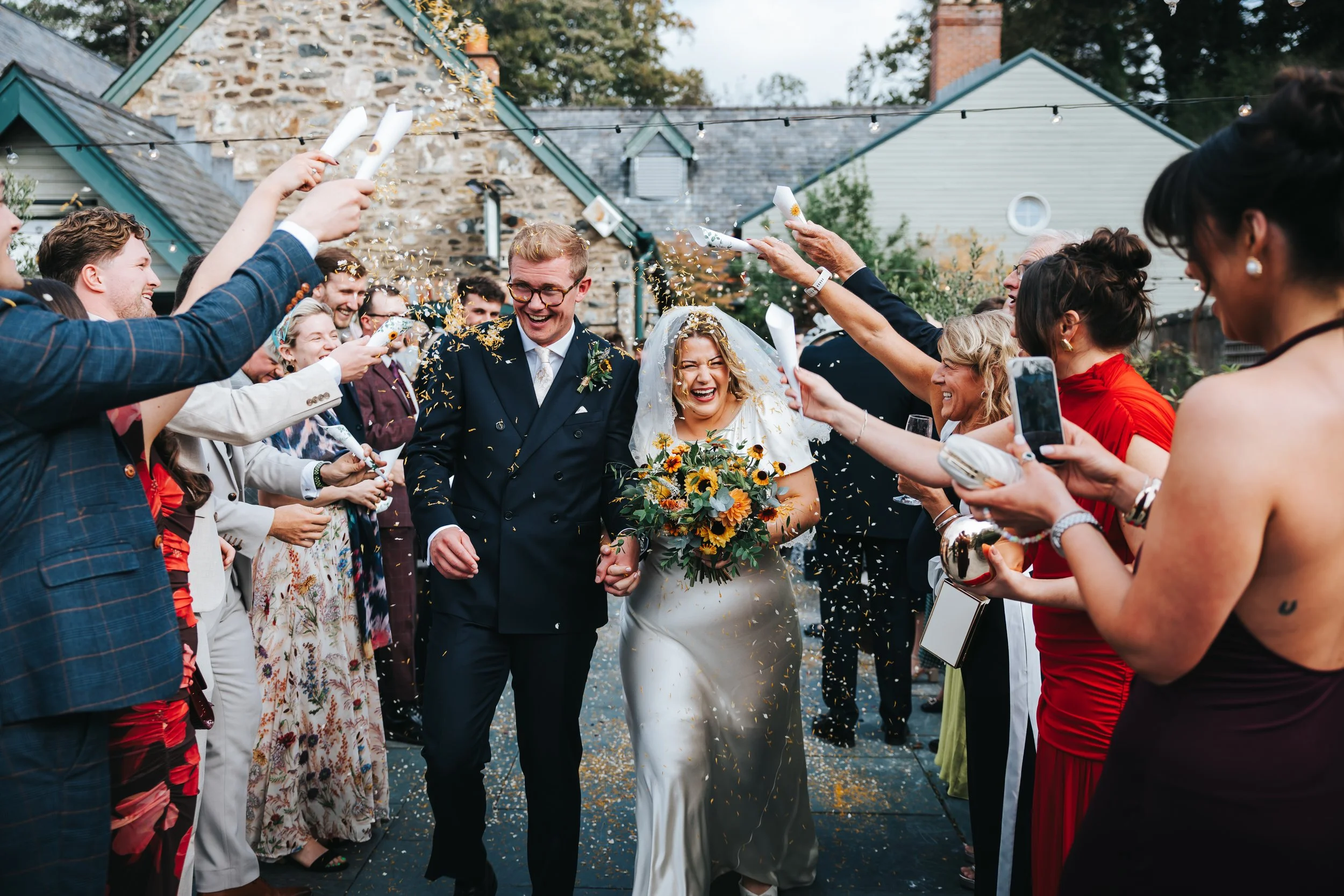 Bride and groom walking hand in hand through a crowd of wedding guests who are throwing confetti and celebrating outside.