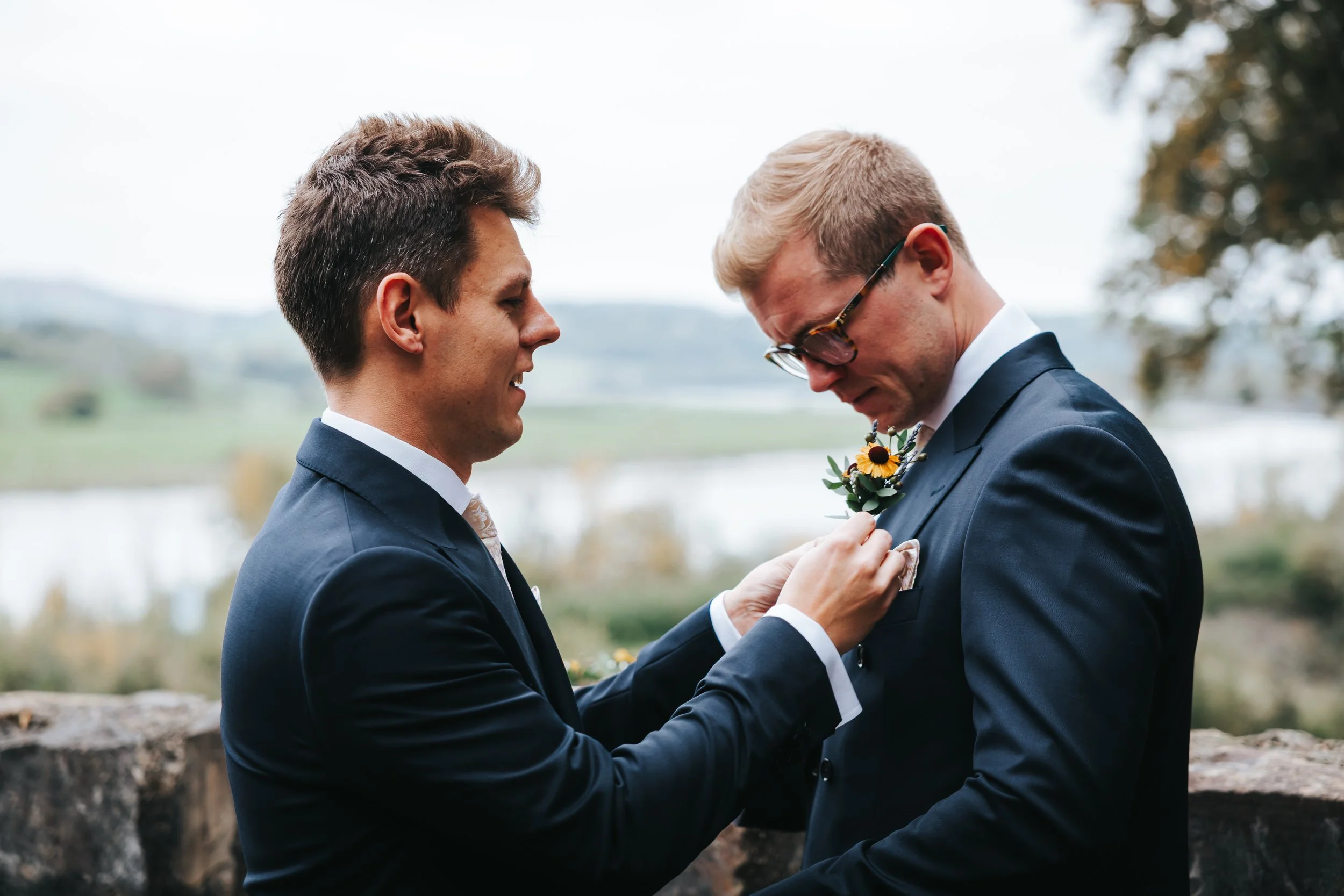 Two men in suits during a wedding, one is pinning a boutonniere on the other's blazer, outdoors with scenic landscape background.