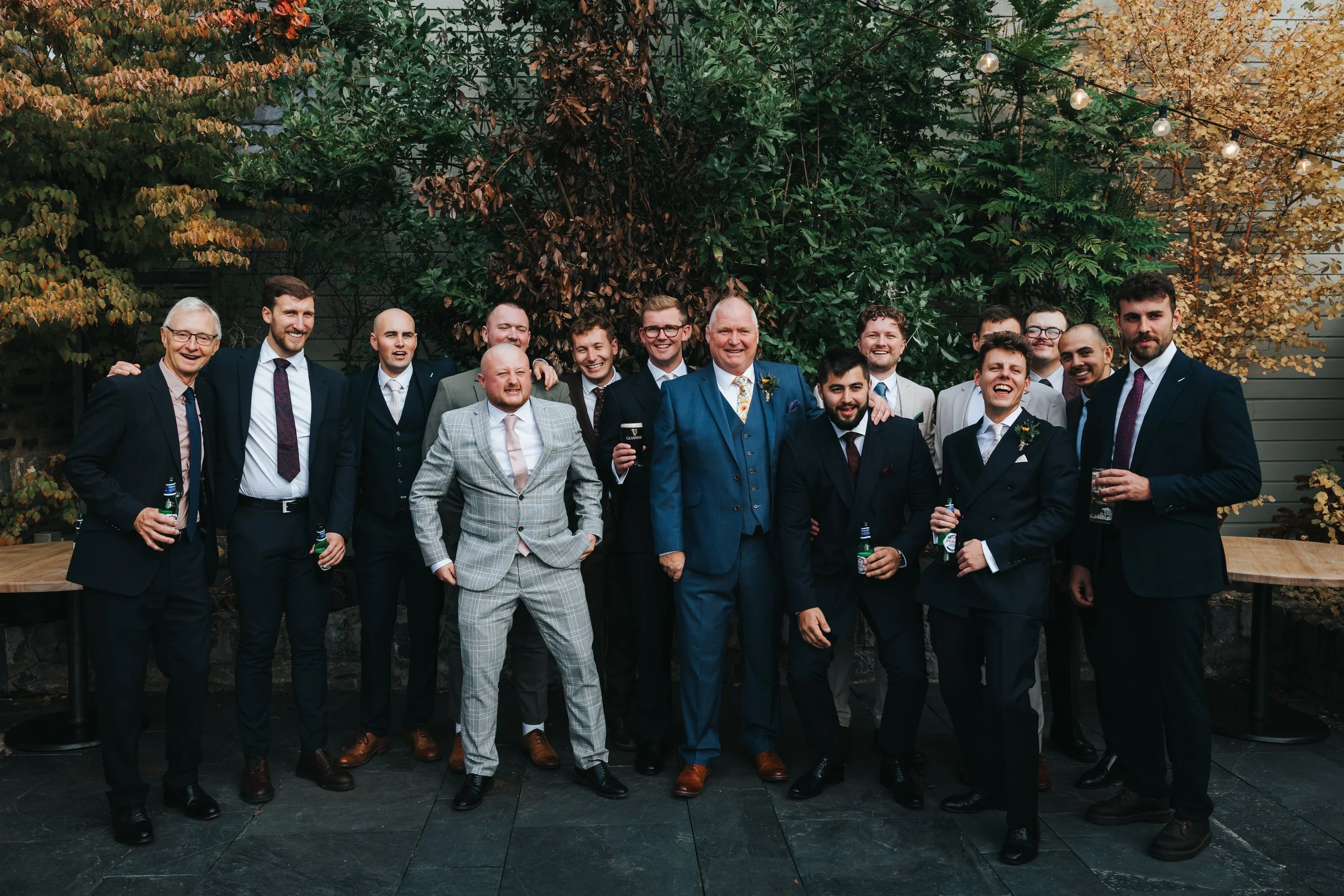 Group of men in suits at a celebration, standing outdoors in front of trees with autumn-colored leaves and string lights.