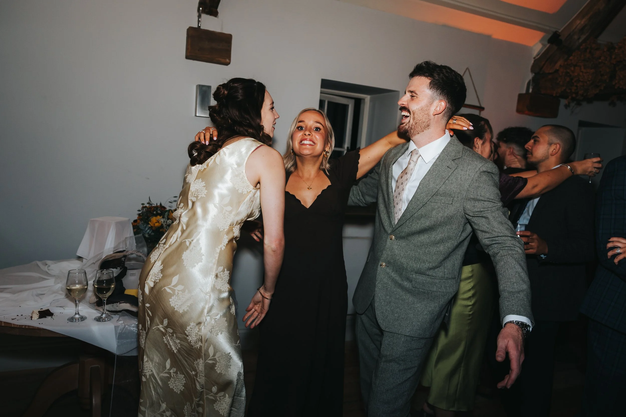 People dancing and celebrating at a party, with two women and a man in the foreground smiling and engaging with each other, surrounded by other guests in the background.