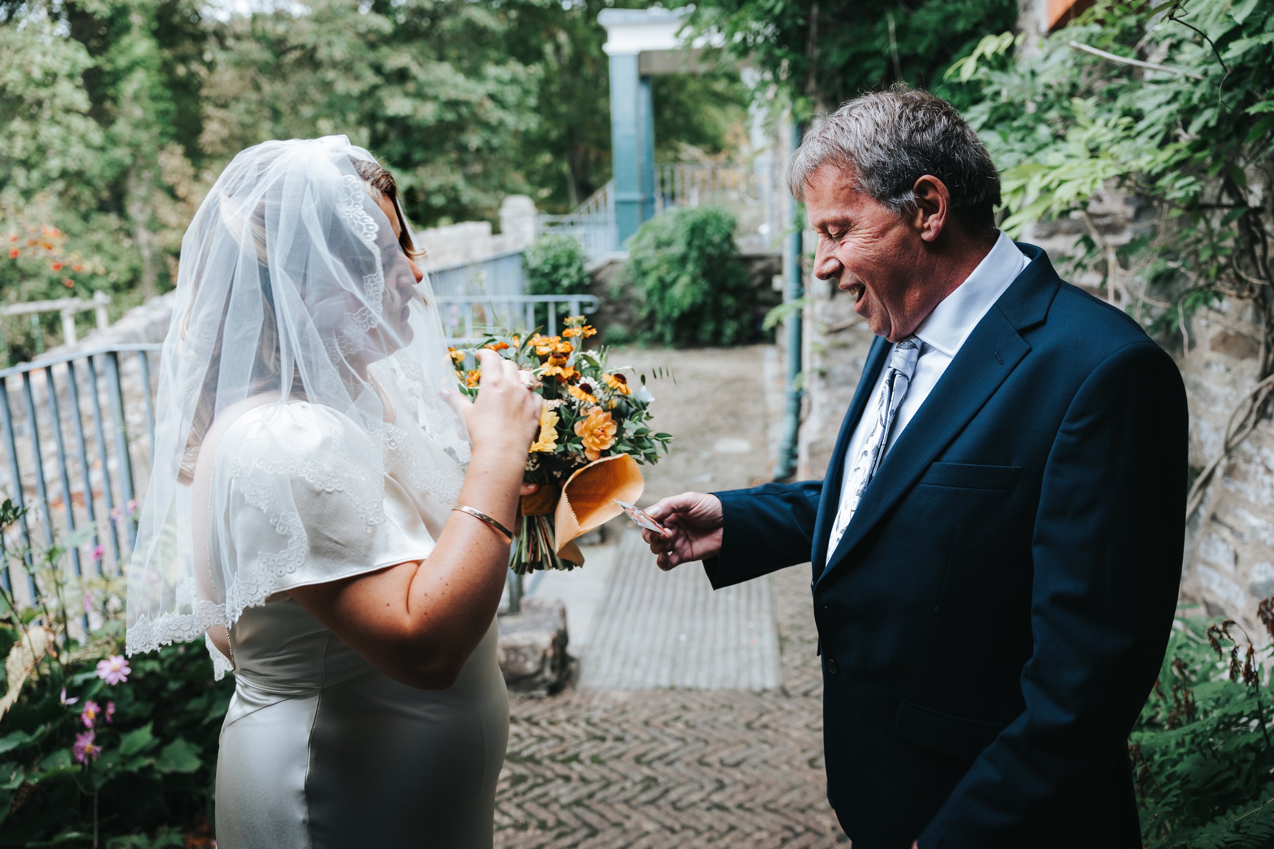 A bride with a veil holding a bouquet of flowers and a man in a suit smiling and showing a photo or card.