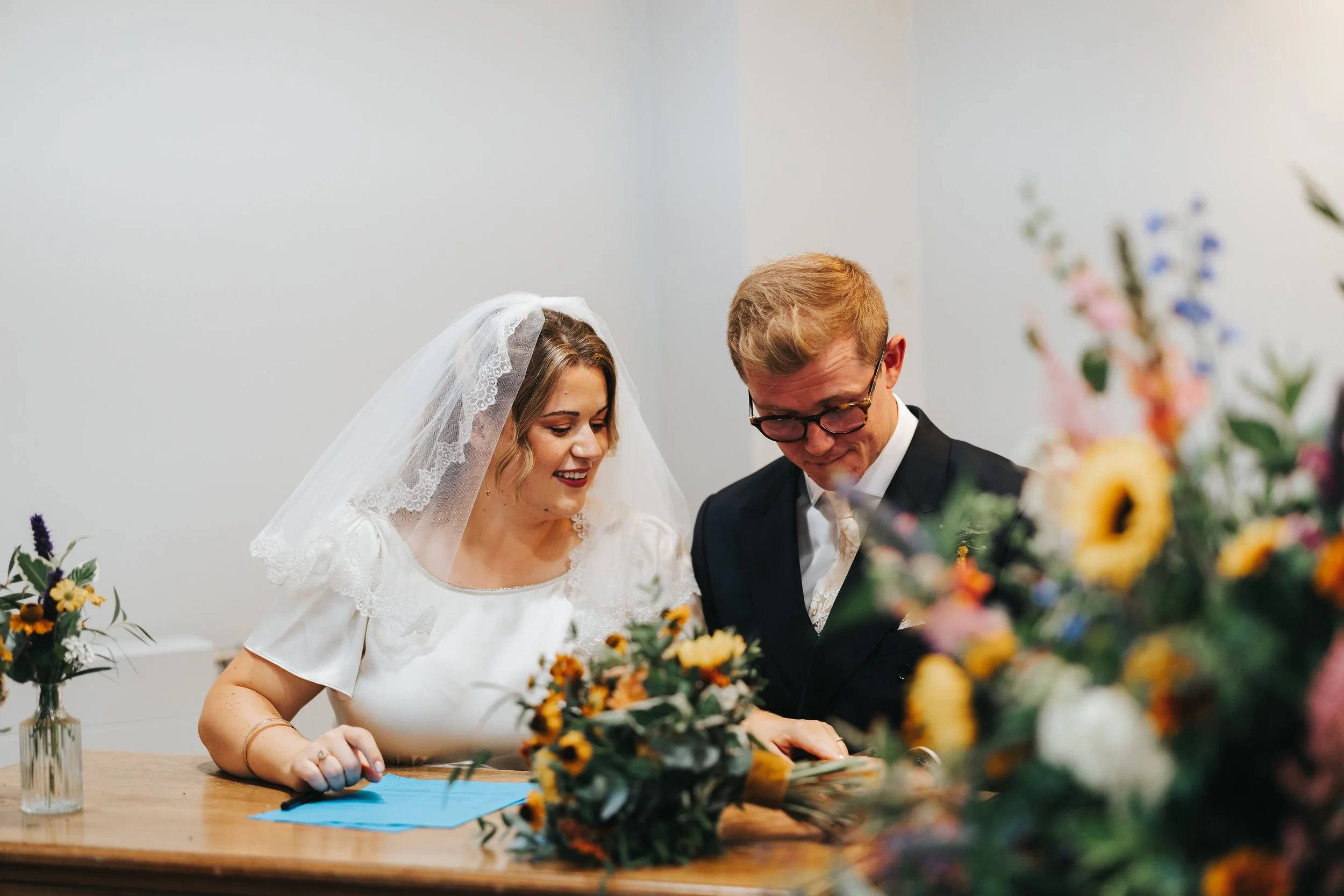 A bride with blonde hair wearing a white dress and veil, and a groom with glasses in a black suit, signing a marriage certificate at a wedding ceremony surrounded by colorful flowers.