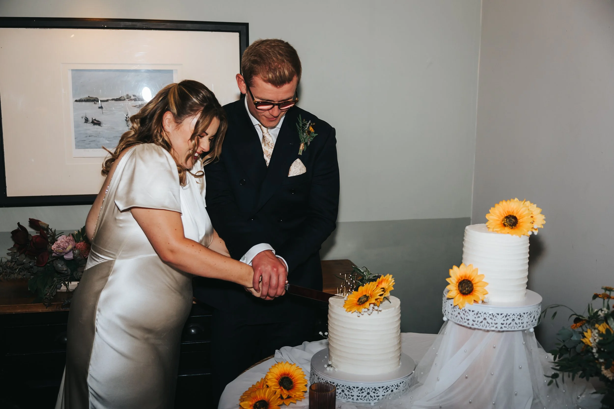 A bride and groom together cutting a wedding cake decorated with sunflowers.