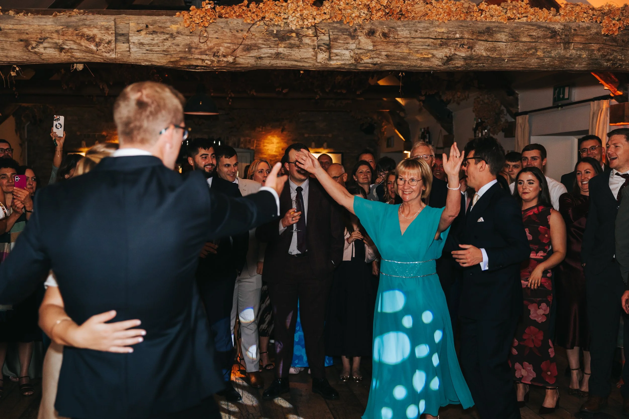 People dancing and enjoying a celebration in a warmly lit indoor venue with exposed brick walls and a wooden beam ceiling.