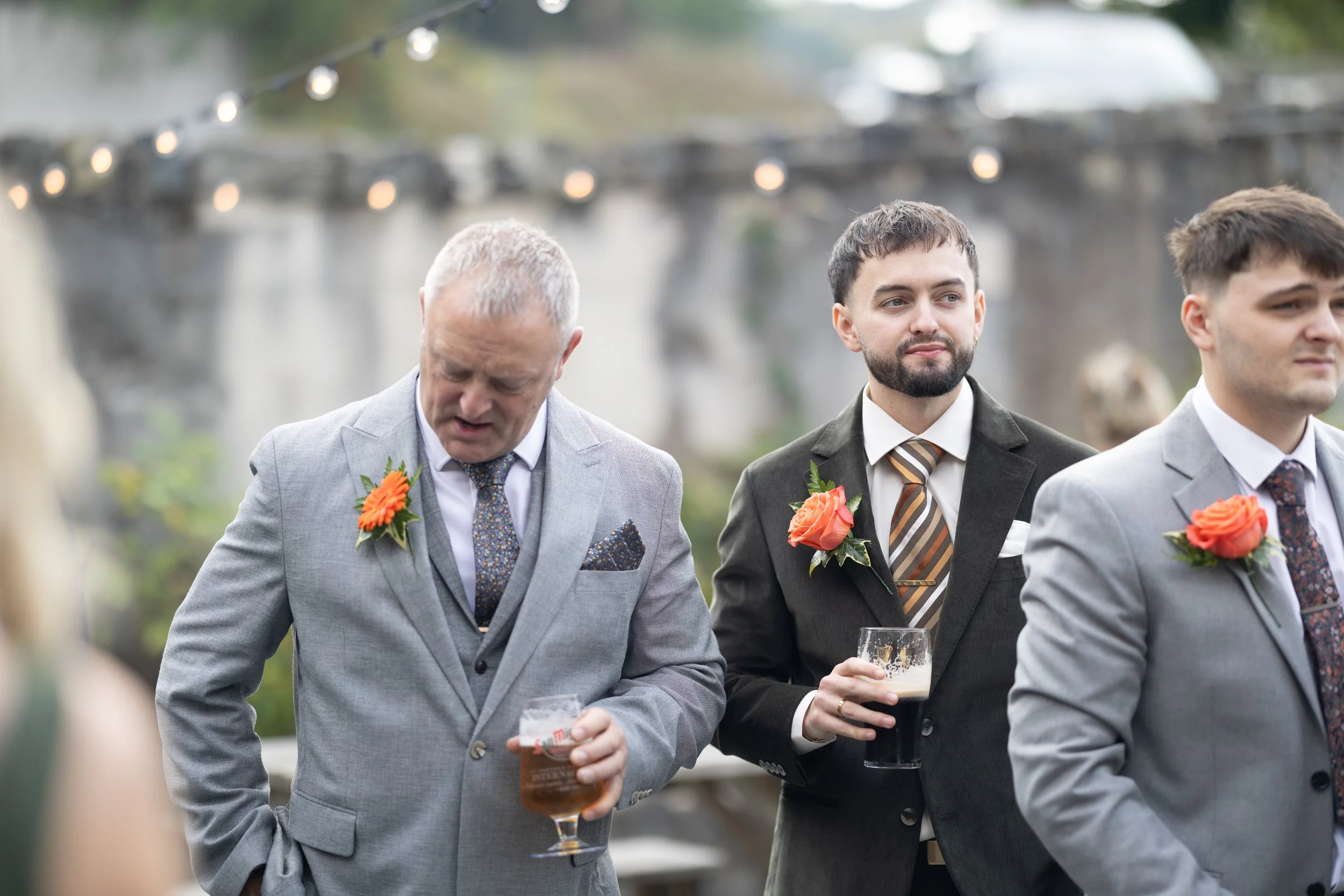 Group of men wearing suits with boutonnières at an outdoor wedding reception, holding drinks.