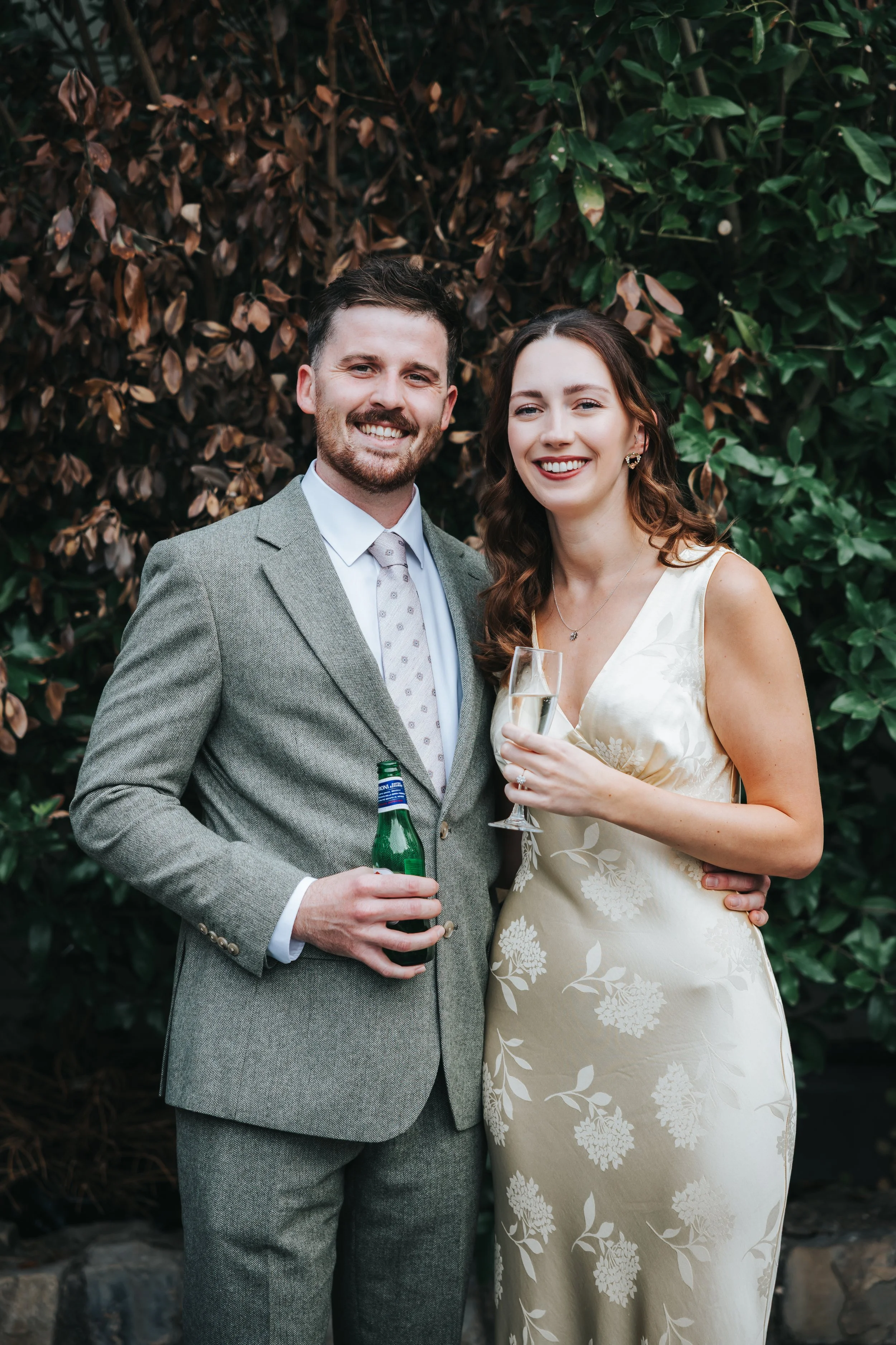 A smiling man and woman in formal attire standing outdoors, holding drinks, in front of green foliage.
