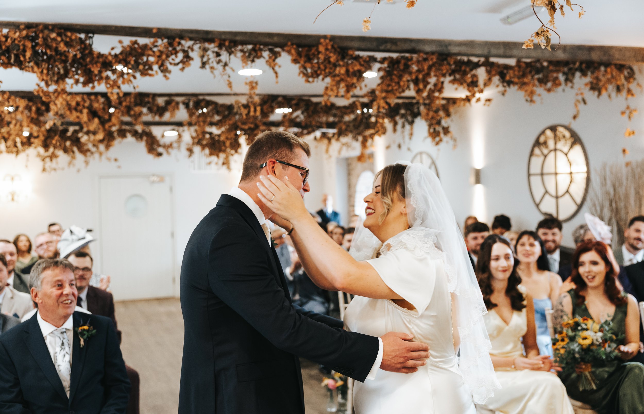 A bride and groom share a kiss during their wedding ceremony in a decorated indoor venue, surrounded by family and friends.