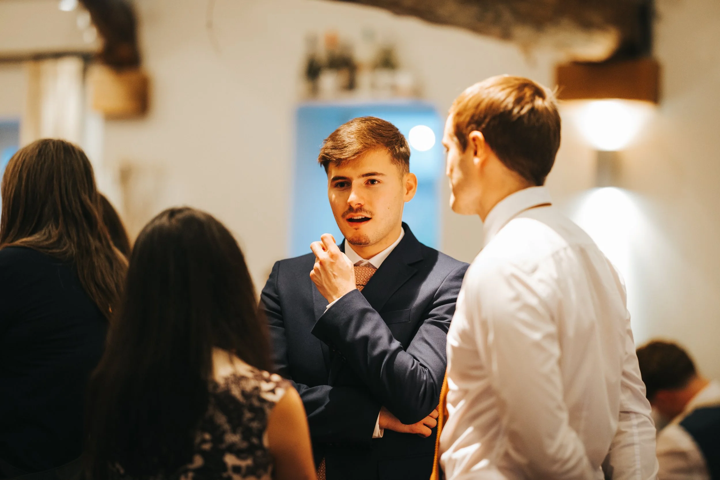 Three people in formal attire having a conversation at an indoor social event, with one man in a black suit and another in a white shirt facing each other while a woman with dark hair and patterned dress listens.