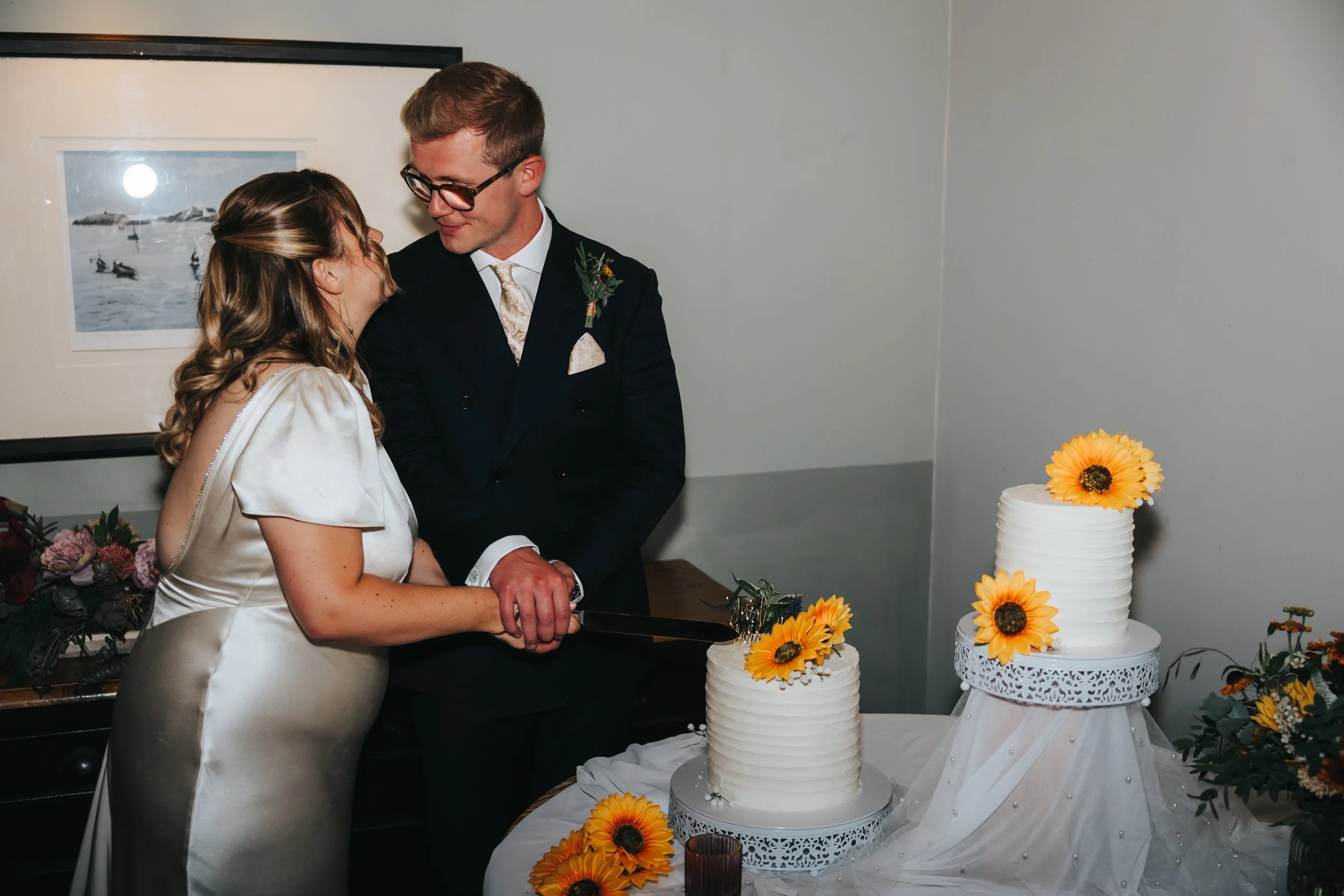 A bride and groom cutting a wedding cake together at their wedding reception, surrounded by sunflower decorations and flowers.