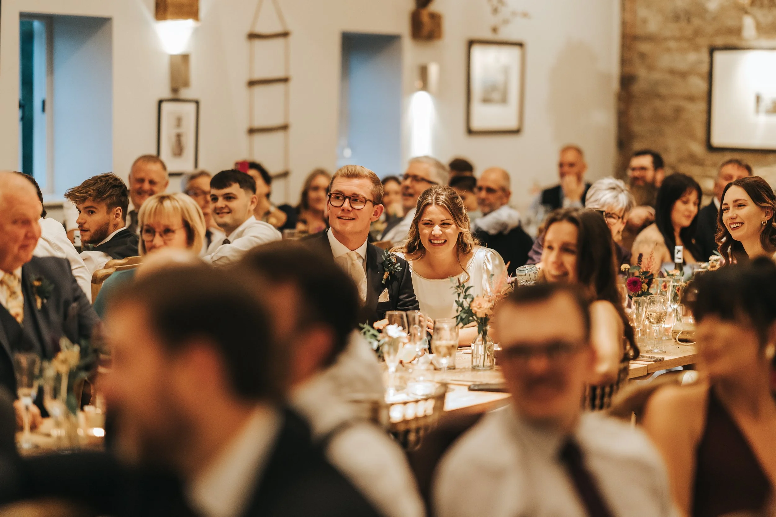 Guests at a reception table smiling and enjoying a celebration, with candles and floral centerpieces, in a warmly decorated venue.