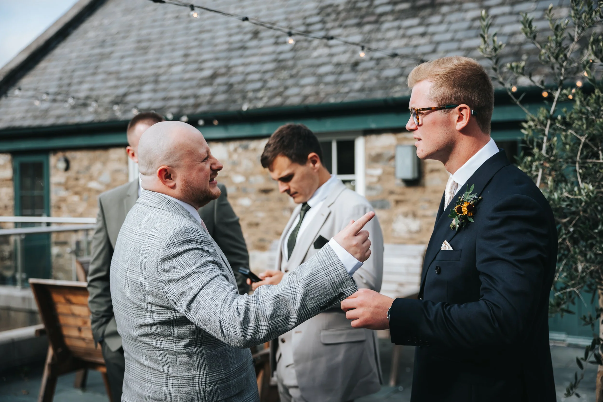 Two men engaged in a conversation at an outdoor wedding or formal event, with one pointing and the other holding a small object, in front of a rustic stone building with string lights overhead.