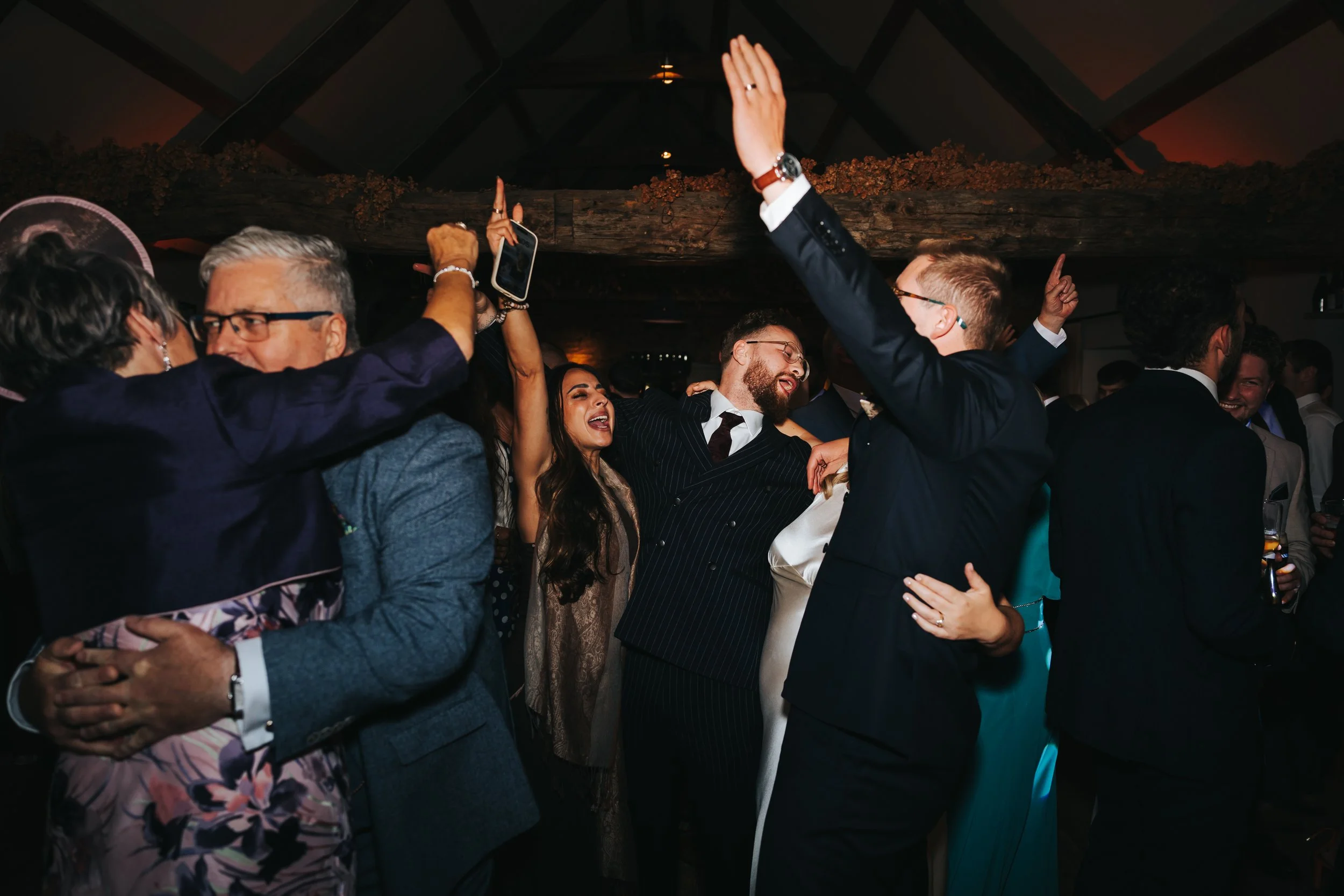 People dancing and celebrating at a party or wedding reception, dressed in formal attire, with some holding drinks, in a dimly lit venue with a rustic ceiling.