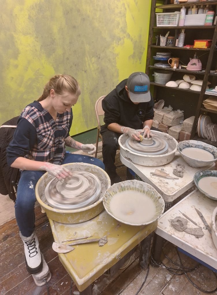 Two people making pottery on electric wheels in a ceramic studio, with brushes and clay tools nearby.