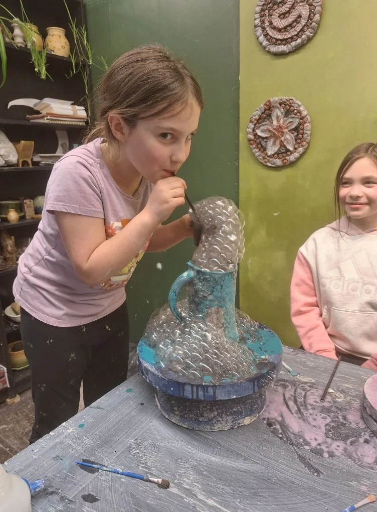A girl with a pink shirt blowing bubbles into a sculpture with a small stick, while another girl watches and smiles. The sculpture is made of glass or similar material and is placed on a table in a craft or art studio.