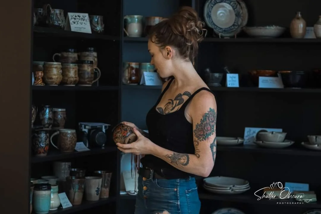 A woman with tattoos on her arms and chest, wearing a black tank top and jeans, is looking at a ceramic bowl while shopping for pottery in a store. She is holding a decorative ceramic item.