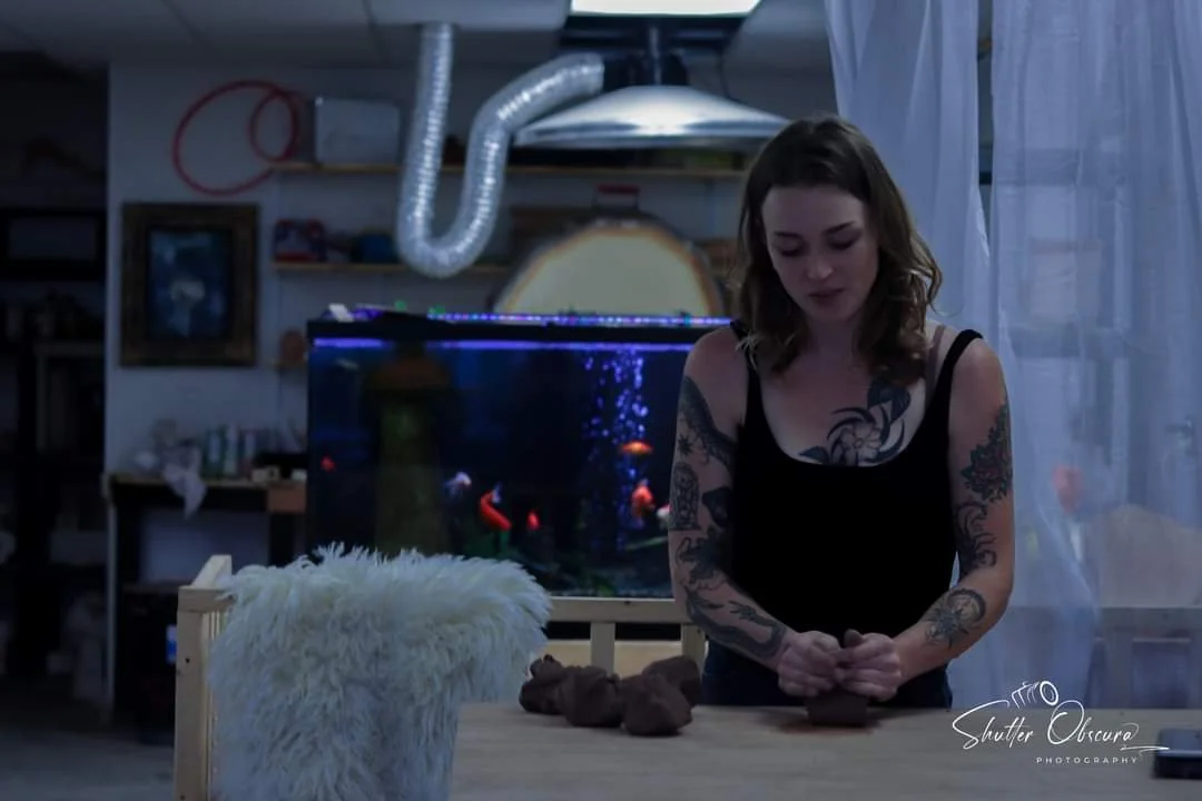 A woman with tattoos on her arms wearing a black tank top, working with clay at a table in a room with a large fish tank in the background.