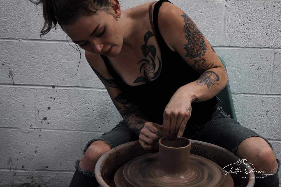 A woman with tattoos on her arms and chest is sitting on a stool, focused on shaping a clay pot on a pottery wheel in a studio with a plain white brick wall.