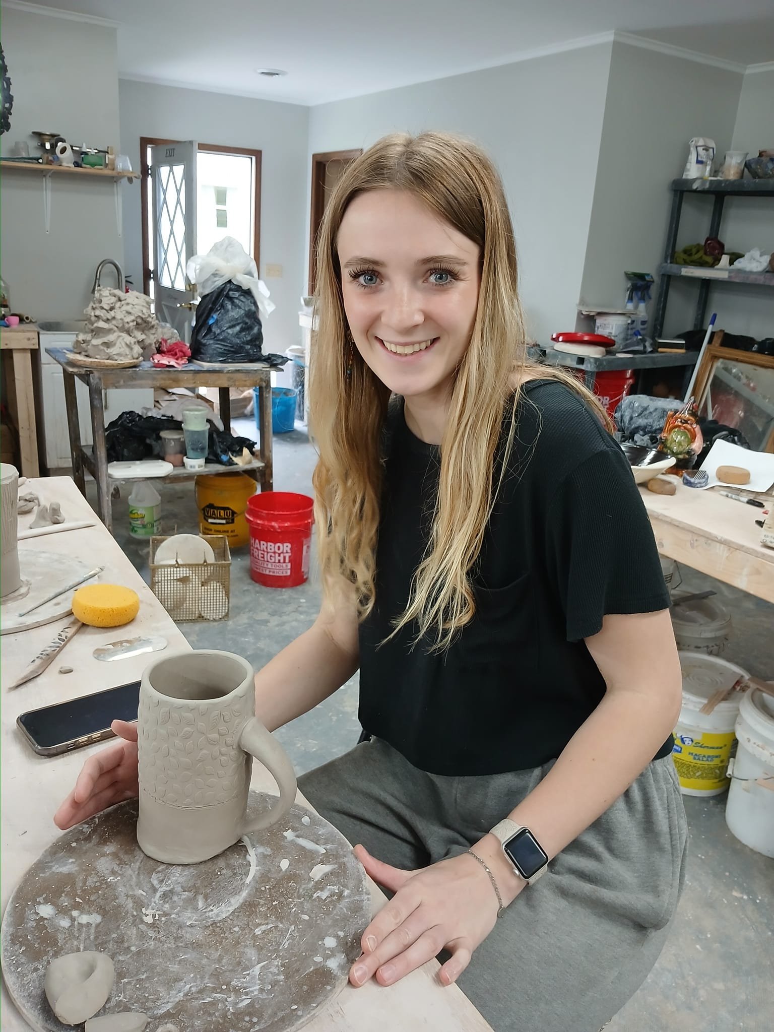 A young woman with long blonde hair smiling and posing with a ceramic mug she created in a pottery studio. The studio is filled with pottery supplies and work stations.