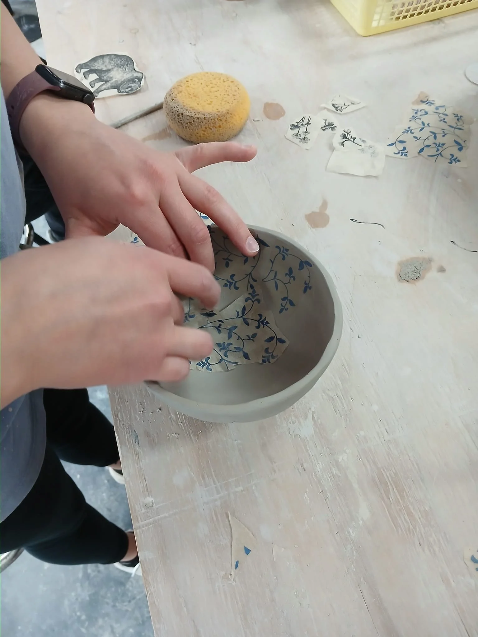 Person applying blue floral transfer to clay bowl on wooden table, with sponge and other transfers nearby.
