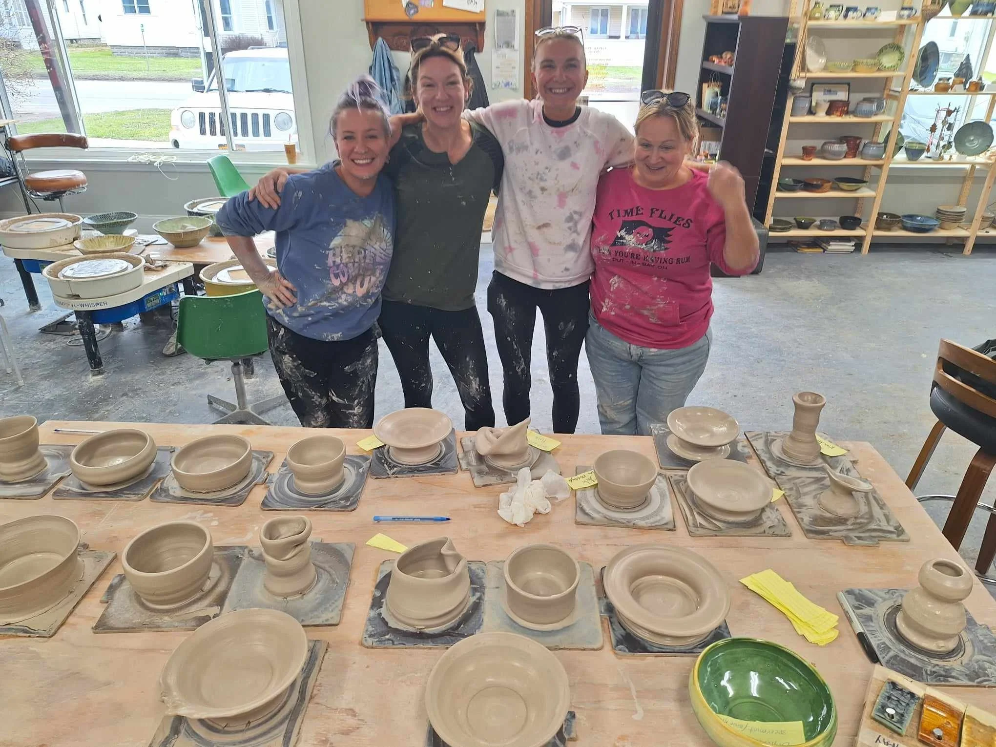 Four women smiling and posing in a pottery studio with pots and bowls on a table in front of them, some covered with plastic and numbered tags, and shelves filled with pottery items in the background.