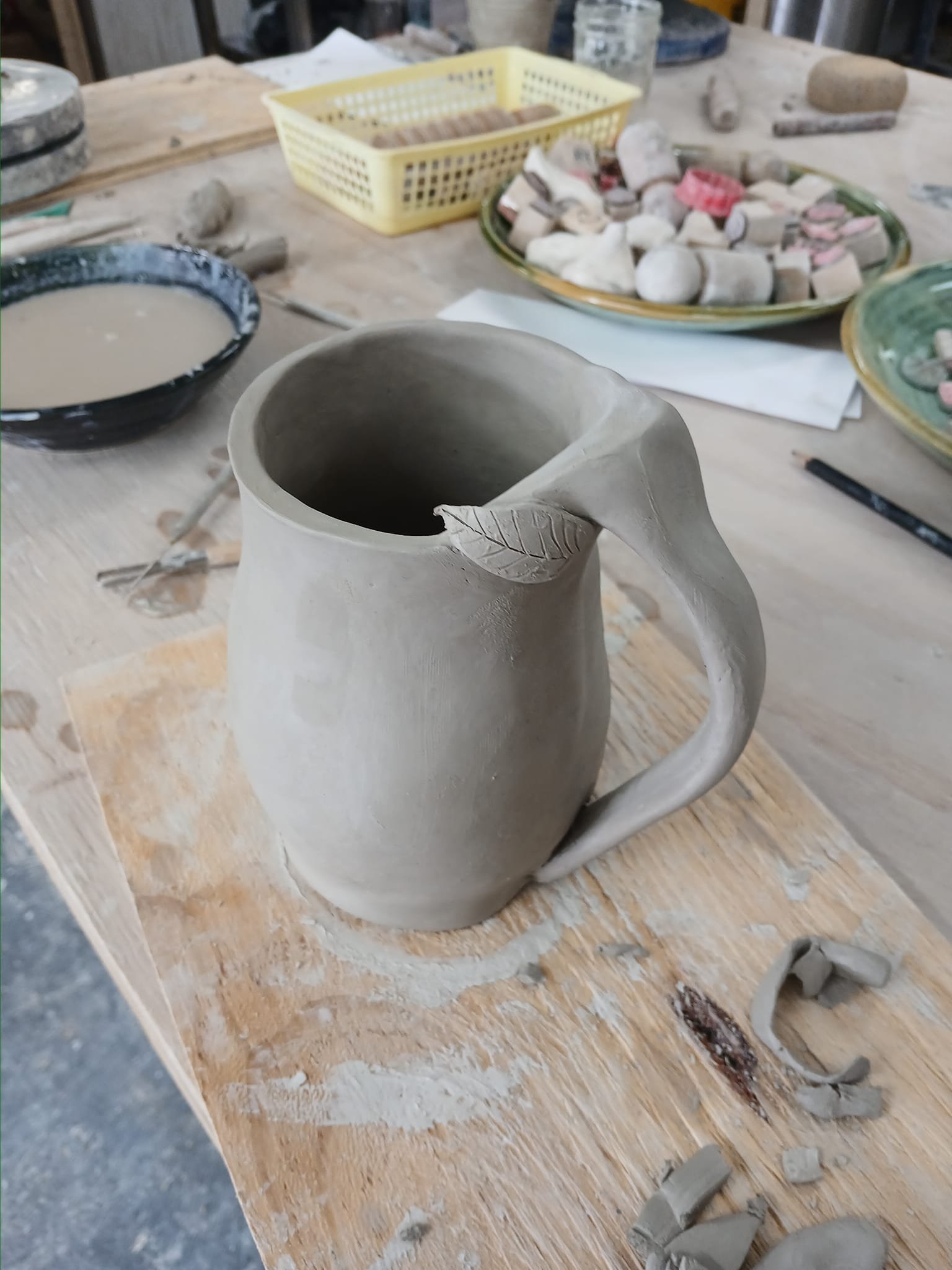 A handmade clay pitcher with a leaf decoration on the handle, on a wooden table with pottery tools and supplies in the background.