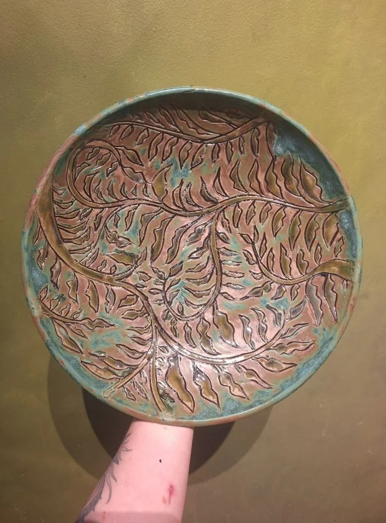 Ceramic plate with leaf and branch engraved design, held by a person with a tattooed arm against a plain background.