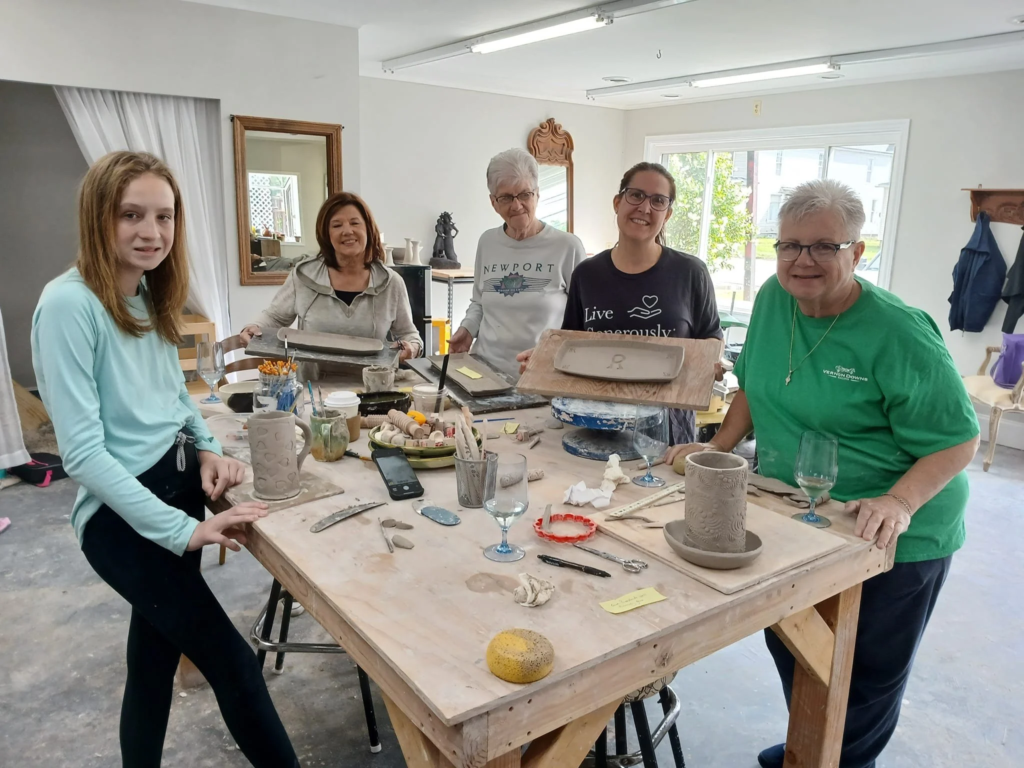 A group of five women in a pottery studio around a worktable, holding clay trays and smiling.