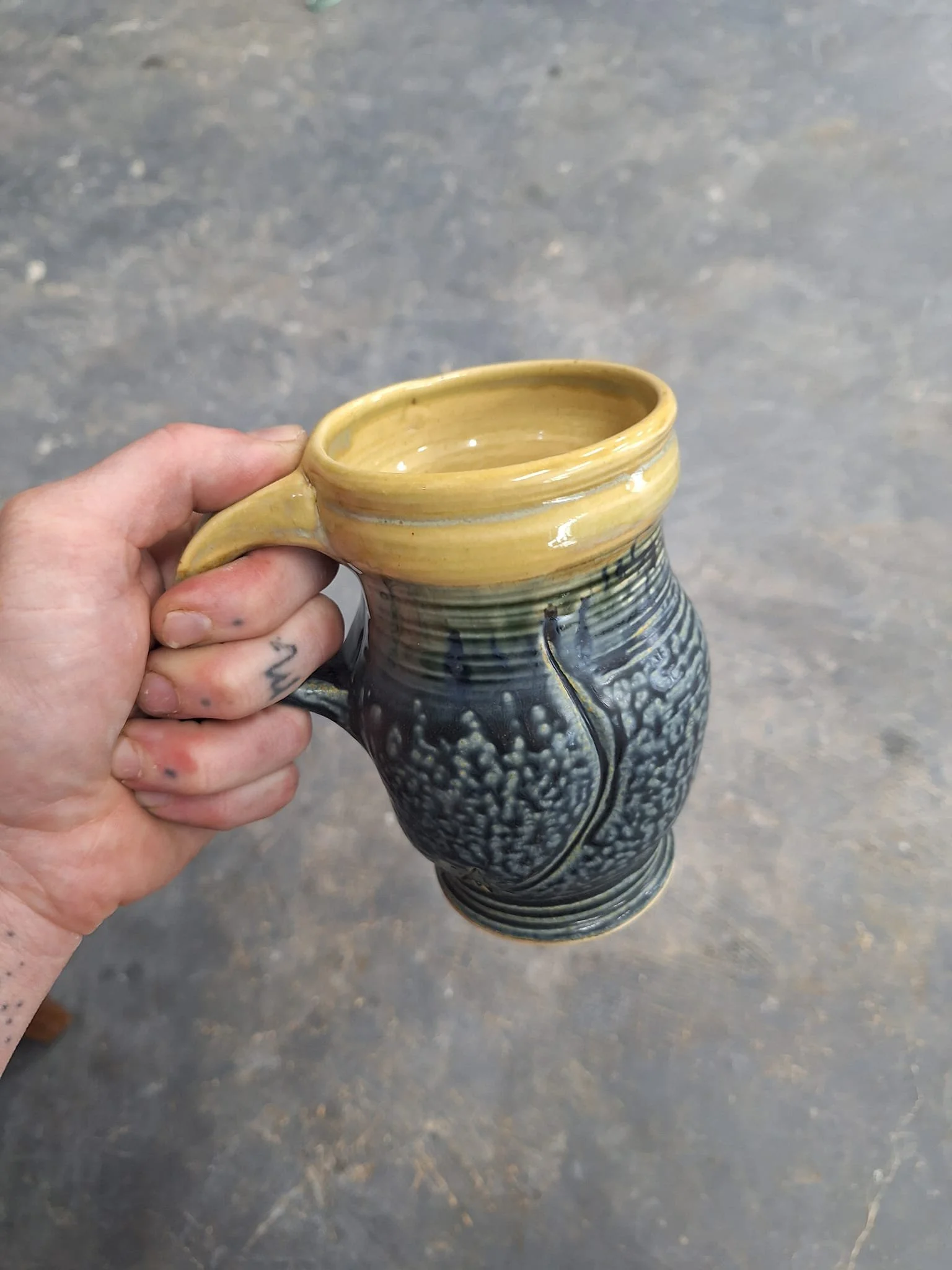 A hand holding a ceramic mug with a yellow interior and a black and gray textured exterior.