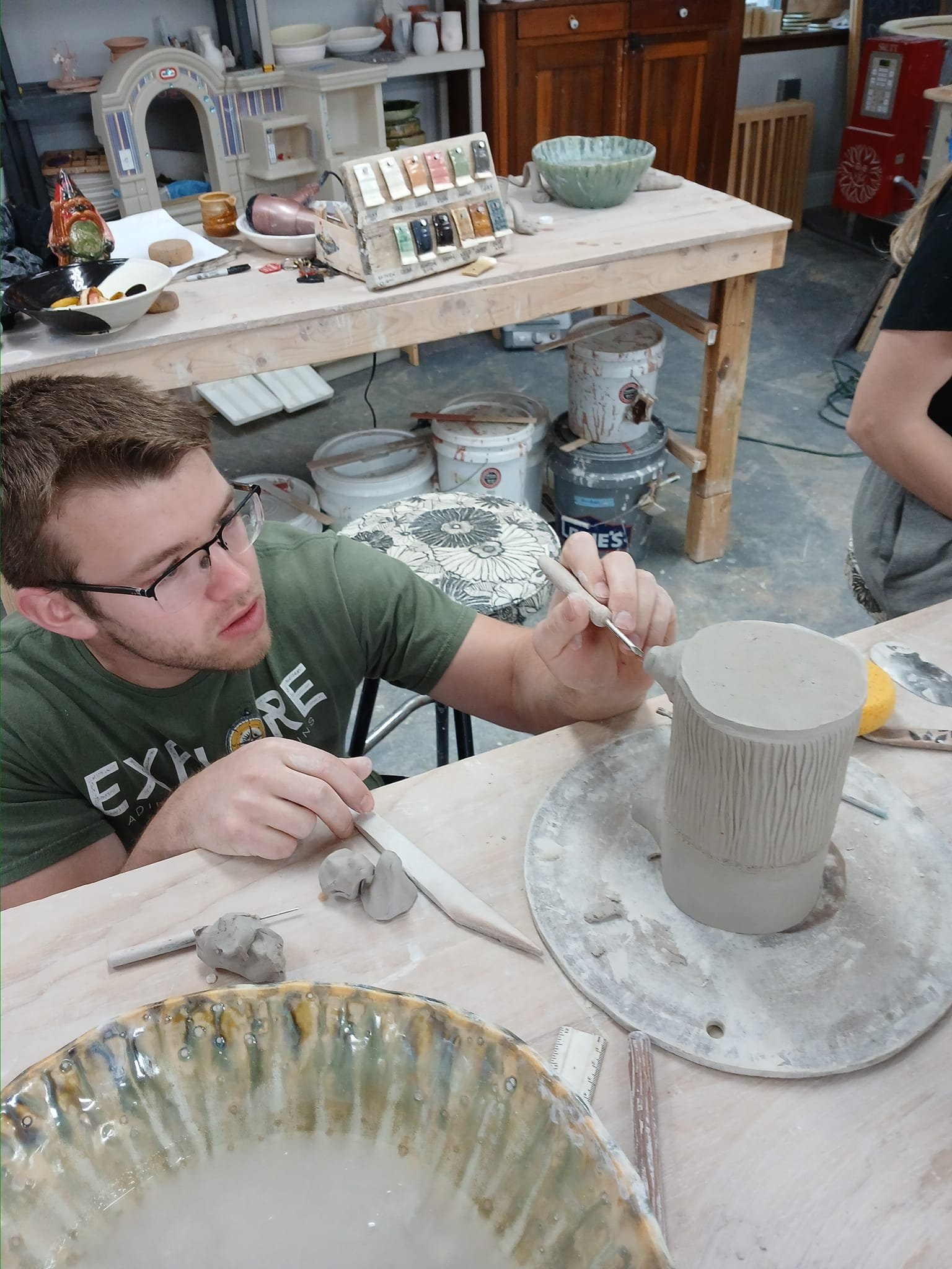 A young man with glasses working on a ceramic pottery piece in a pottery workshop, focusing on carving or detailing a vertical cylindrical vessel.