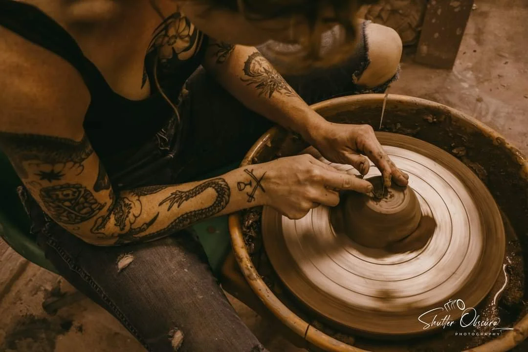 A person with tattoos on both arms is shaping wet clay on a pottery wheel to make a ceramic pot.