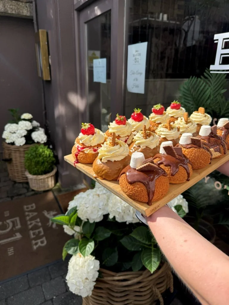 A wooden tray of assorted decorated cream-filled pastries topped with strawberries, marshmallows, and cookies, held by a person outside a bakery.