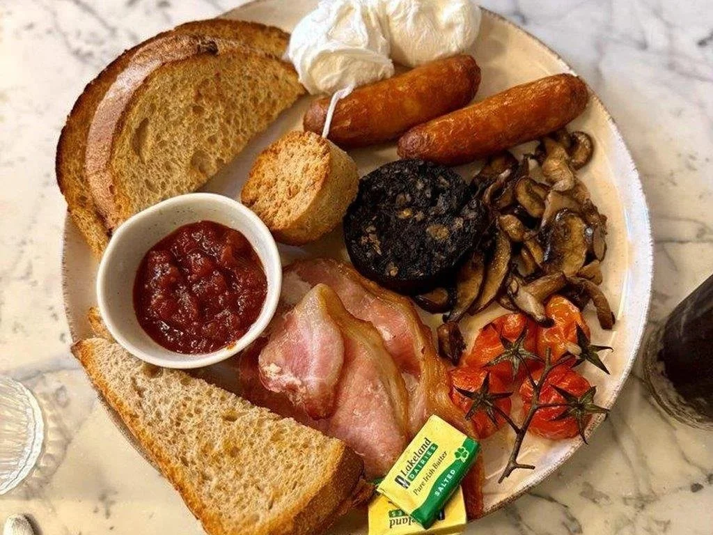 Breakfast platter with toasted bread, fried eggs, sausage links, bacon, grilled tomatoes, sautéed mushrooms, black pudding, a small bowl of tomato relish, and butter.