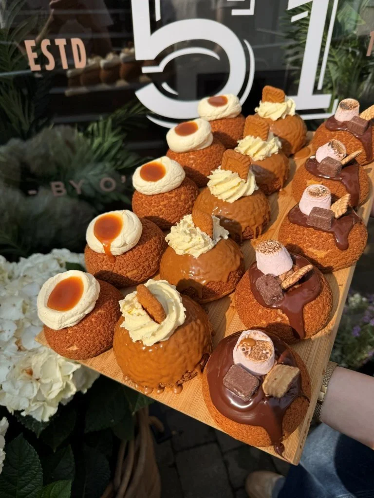 Assorted desserts on a wooden tray, including profiteroles topped with cream and caramel, and chocolate éclairs with various toppings, displayed in front of a black sign with white text.