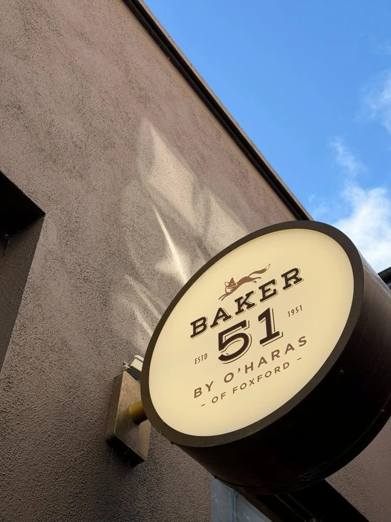 Circular cafe sign reading "Baker 51 by O'Hara's of Foxford," with an illustration of a cat, mounted on an exterior gray wall, with blue sky and clouds in the background.