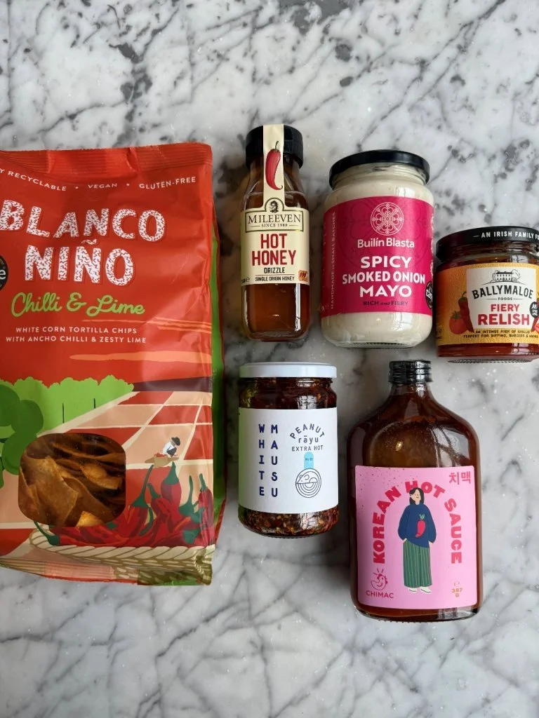 Collection of various condiments and snacks on a marble surface, including a bag of Blanco Nino chili and lime potato chips, a bottle of Mileven hot honey, a jar of spicy smoked onion mayo, a jar of Ballymaloe fiery relish, a jar of white and purple 
