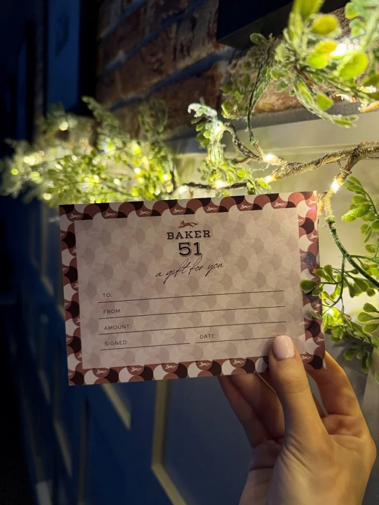 A gift card from Baker 51, labeled 'a gift for you' with fields for To, From, Amount, Signed, and Date, held in front of a plant with green leaves and small yellow flowers.