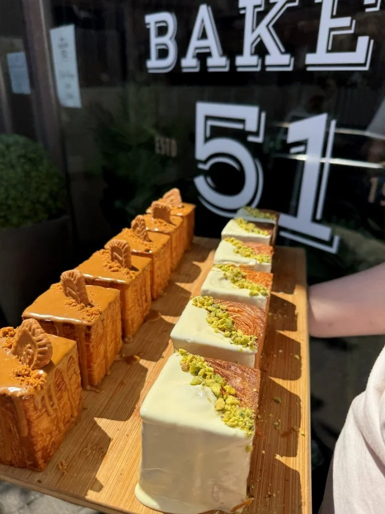 Assorted rectangular cakes placed on a wooden serving board, with a bakery sign in the background.