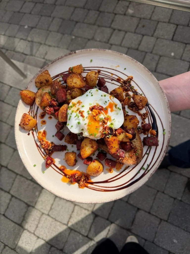 Plate of roasted potatoes topped with a poached egg, garnished with chopped chives and drizzled with balsamic glaze and hot sauce, on a tiled sidewalk.
