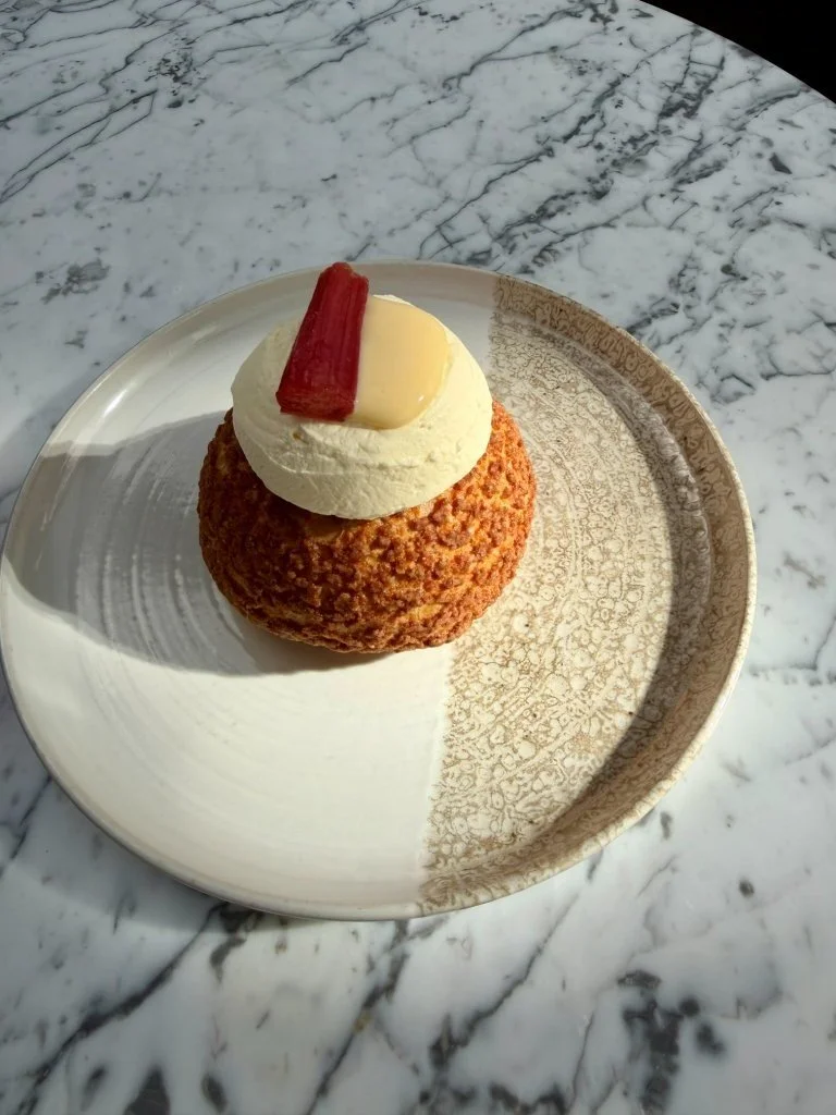 A dessert with a round, golden-brown crumb coating, topped with whipped cream, a yellow candy piece, and a red candy strip, served on a decorative beige and white plate on a marble surface.