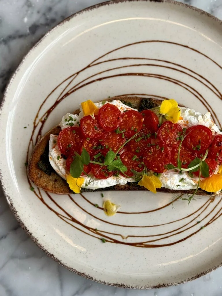 A piece of toasted bread topped with whipped cheese, cherry tomatoes, microgreens, and edible yellow flowers, garnished with a drizzle of balsamic glaze on a white plate with brown decorative lines.