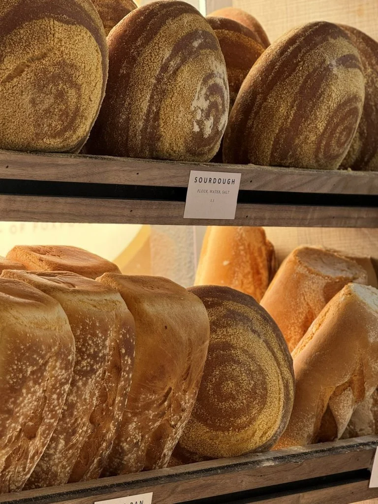 Loaves of sourdough bread on wooden shelves with a label that reads "Sourdough, flour, water, salt, $5.5"