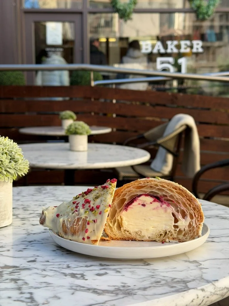 A plate with a slice of cake and a croissant on a marble table outside a bakery café. The bakery's name sign reads 'BAKER 51' in the background.