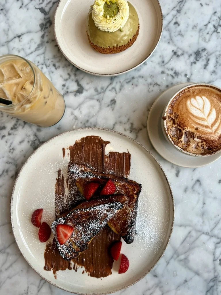 A marble table with three desserts: a green dome-shaped cake decorated with white frosting and pistachios, a slice of French toast with strawberries and powdered sugar, and a chocolate croissant with chocolate shavings, accompanied by an iced coffee 