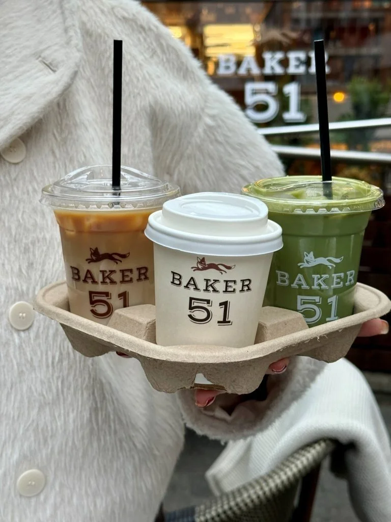 Hand holding a tray with three drinks from Baker 51, one iced coffee, one hot coffee, and one green smoothie, in a cafe setting.
