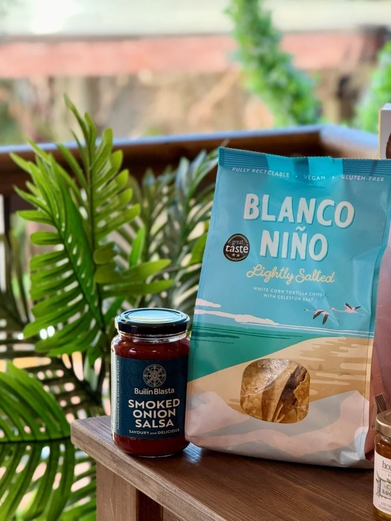 A jar of smoked onion salsa and a bag of Blaco Niño lightly salted white corn tortilla chips on a wooden surface with green plants in the background.