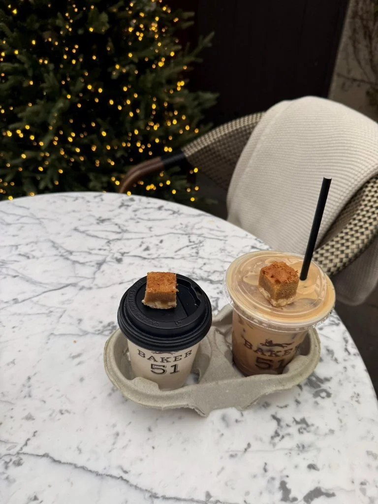 Two beverages with cookies on top sitting on a marble table near a Christmas tree with lights.
