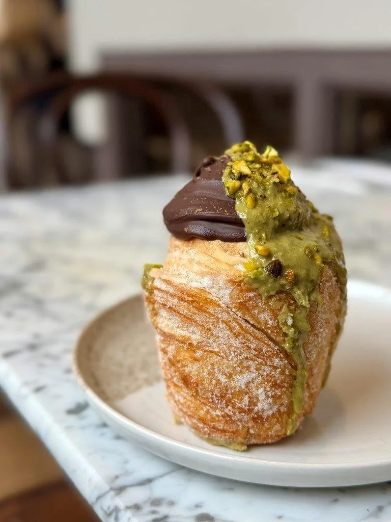 A close-up of a dessert pastry topped with chocolate and pistachios on a white plate, on a marble table.