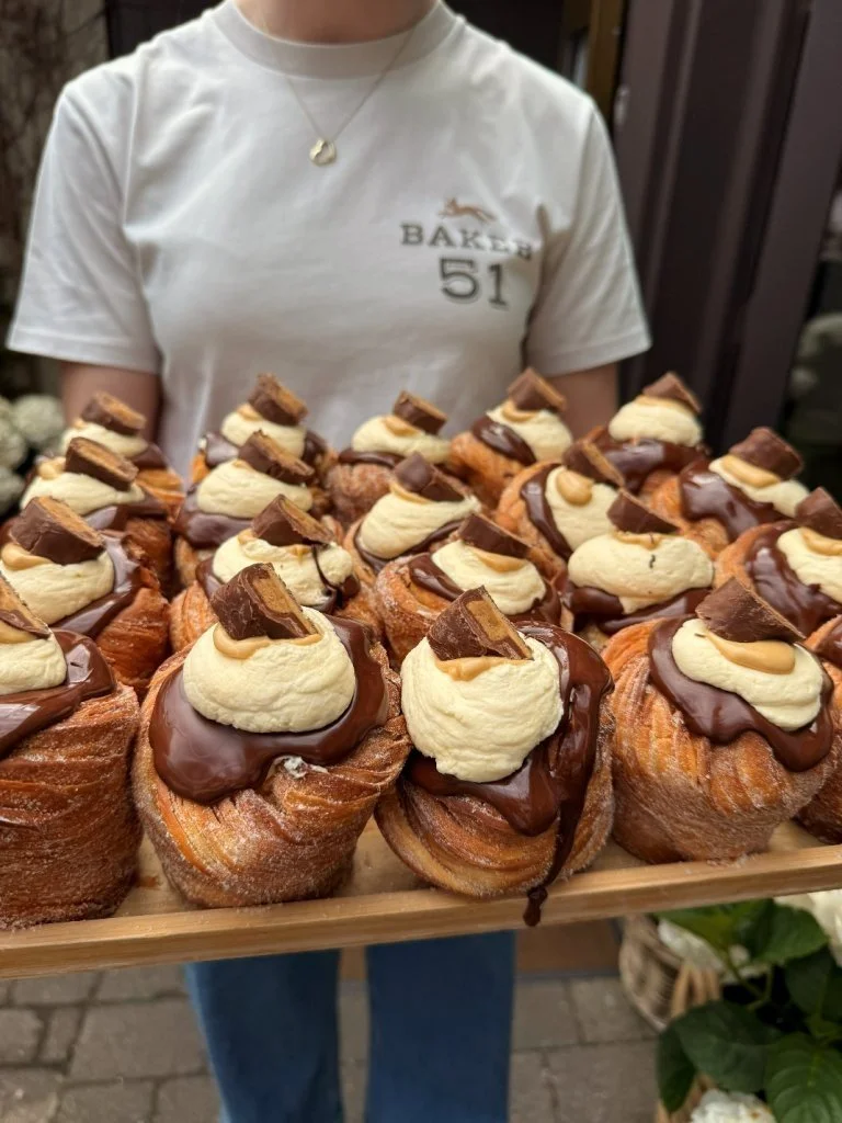 Person holding a wooden tray filled with multiple caramel cream puffs topped with whipped cream, chocolate drizzle, and small pieces of chocolate.