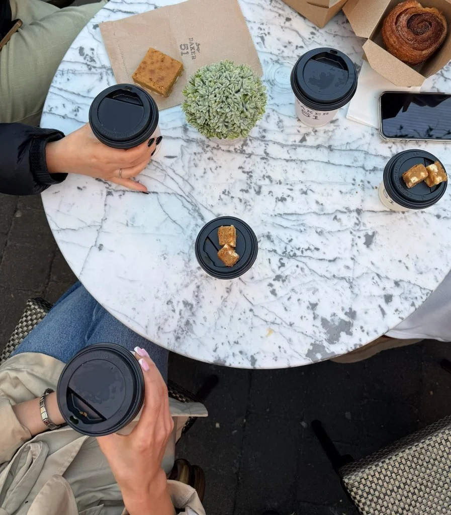 A top-down view of a round marble table with six coffee cups, some with caramel pieces on lids, a green plant, a pastry, a cinnamon roll, a smartphone, and a sandwich wrapped in paper, with people holding coffee cups.