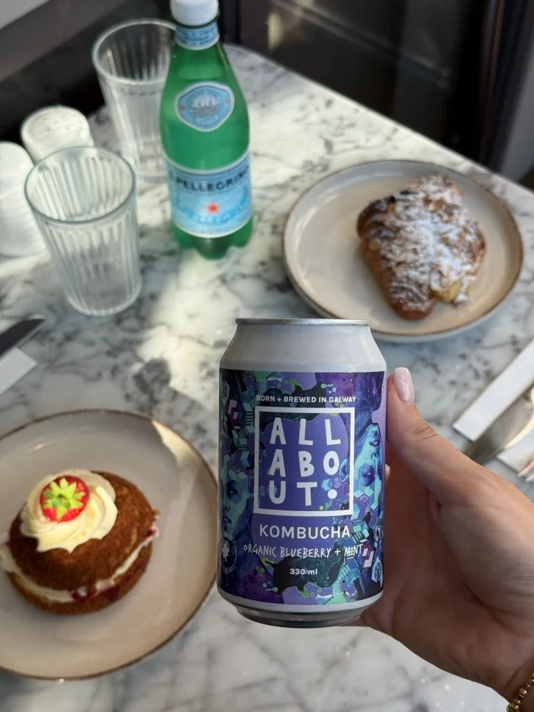 Hand holding a can of All About Kombucha organic blueberry mint, with a marble tabletop featuring two plates of desserts, a bottle of sparkling water, and two empty glasses.